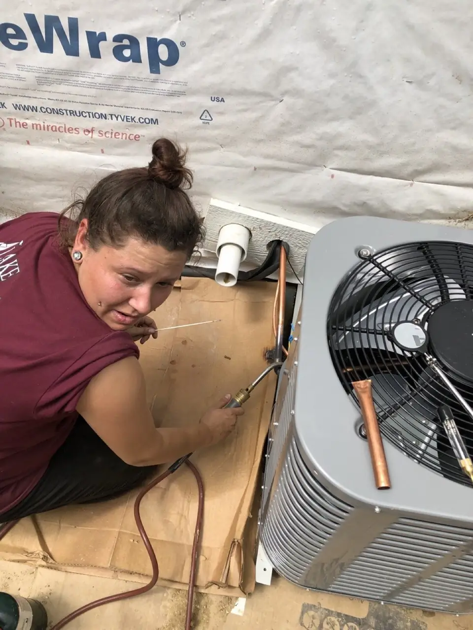 Technician crouched beside an outdoor condensing unit, brazing the copper refrigerant lines with a torch